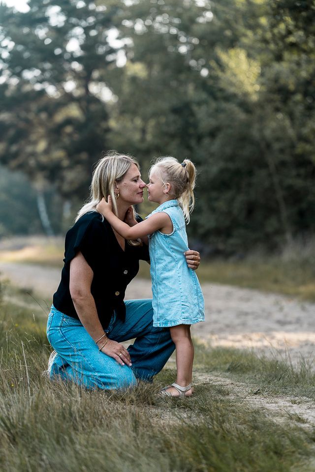 Een vrouw knielt neer en omhelst en kust een jong kind buiten op een zandpad tussen de bomen.