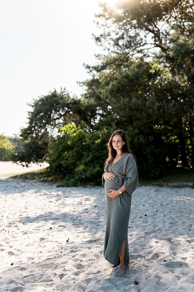 Een zwangere vrouw in een groene jurk ligt op een zandstrand en houdt haar buik vast. Het zonlicht filtert door de bomen.