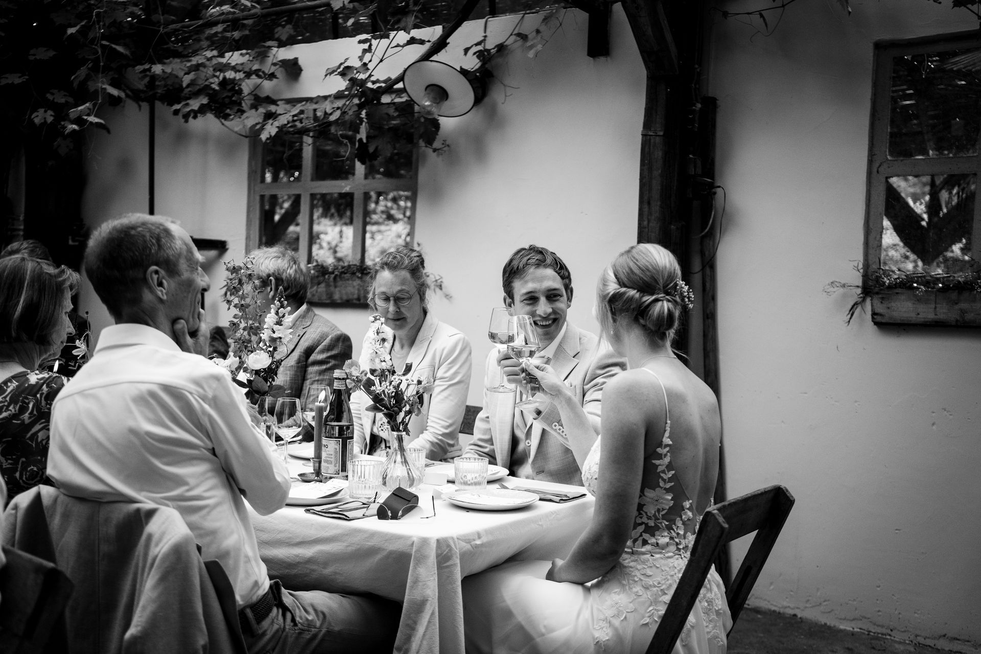 Bruiloftsreceptie, mensen zitten aan een tafel. Een vrouw heft een glas, anderen glimlachen, onder een met wijnranken begroeide pergola.