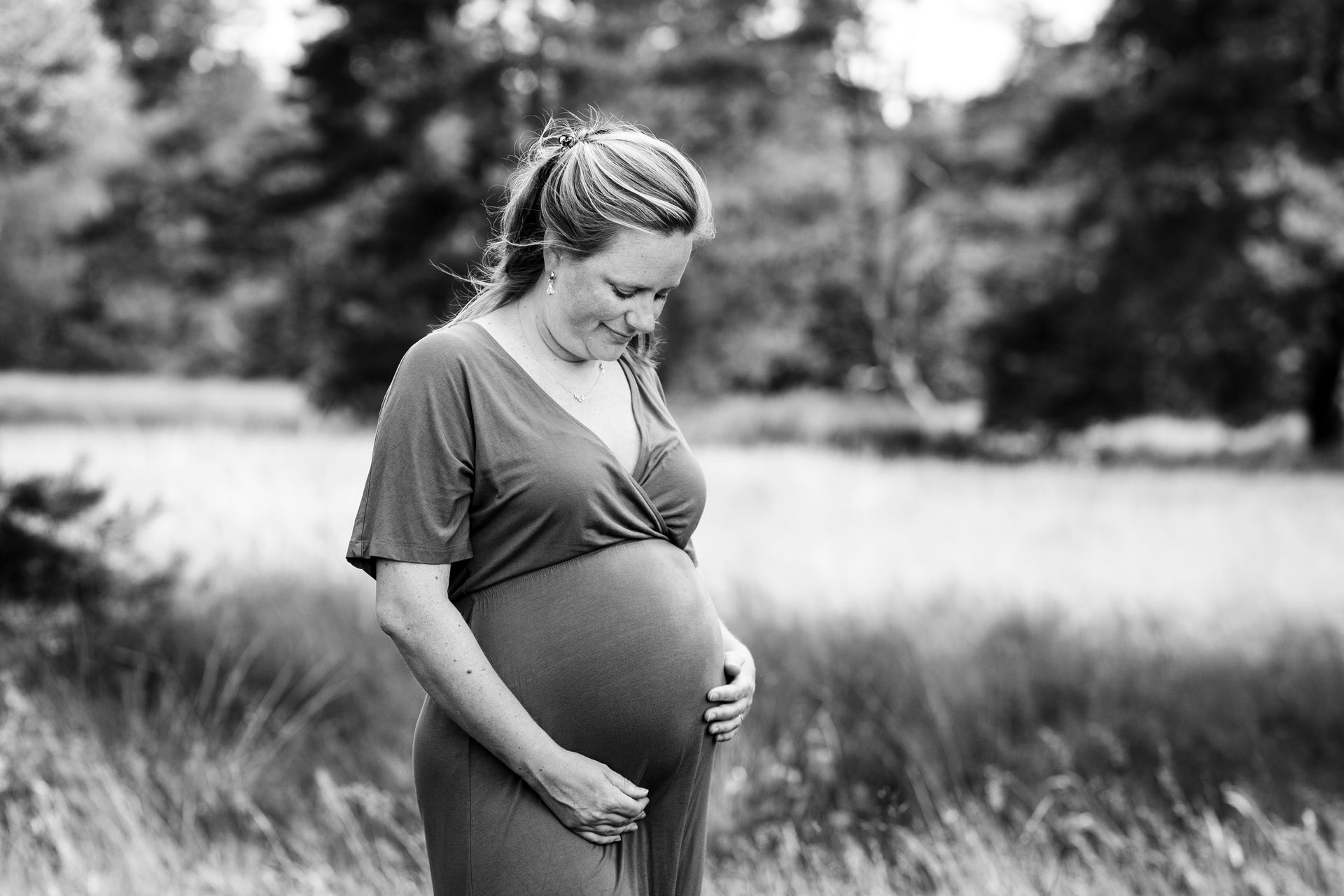 Een zwangere vrouw glimlacht terwijl ze haar buik vasthoudt in een grasveld. Zwart-witfoto, buitenopname.