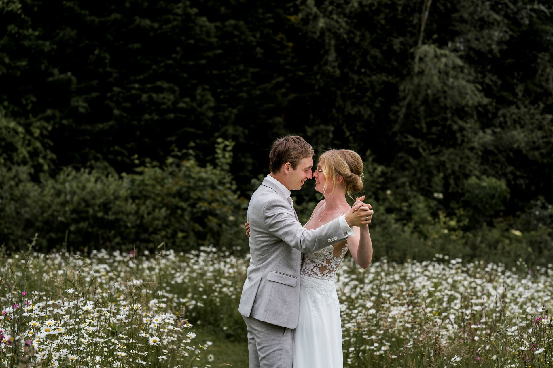 Een paar danst in een veld met witte bloemen. De man draagt een grijs pak en de vrouw een trouwjurk; ze staan dicht bij elkaar.