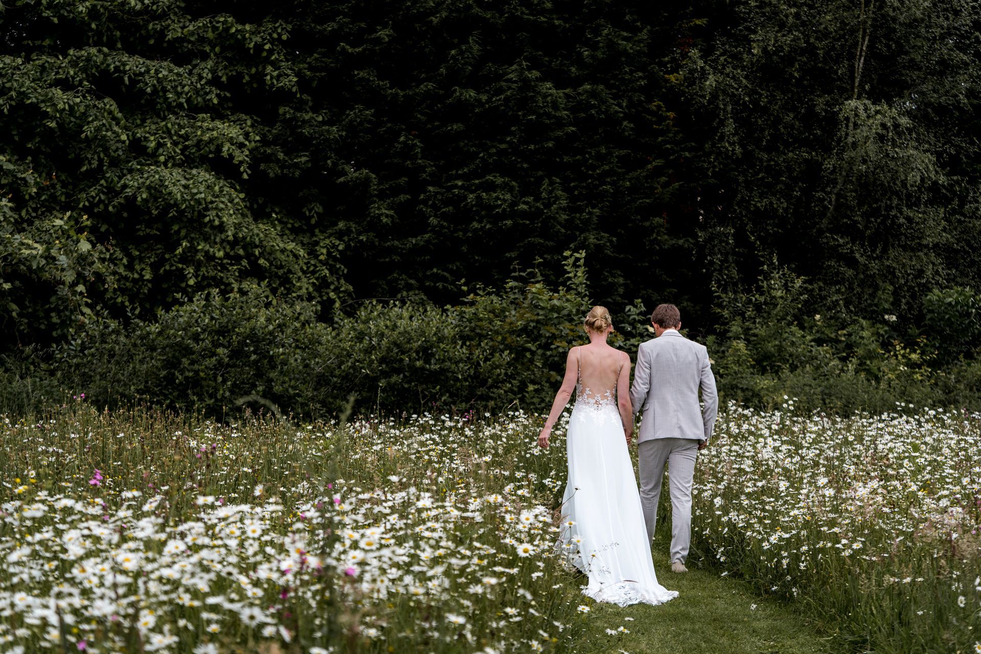 Een stel loopt hand in hand door een veld vol wilde bloemen richting een donker bos. De bruid draagt ​​een witte jurk.