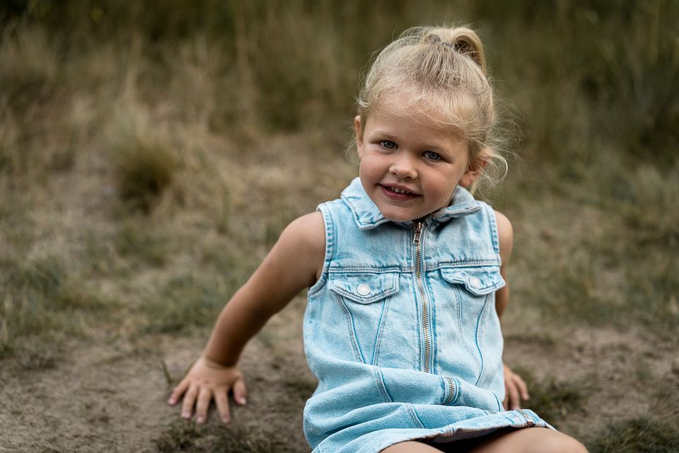 Een blond kind in een spijkerjurkje lacht terwijl ze buiten op de aarde en het gras zit.