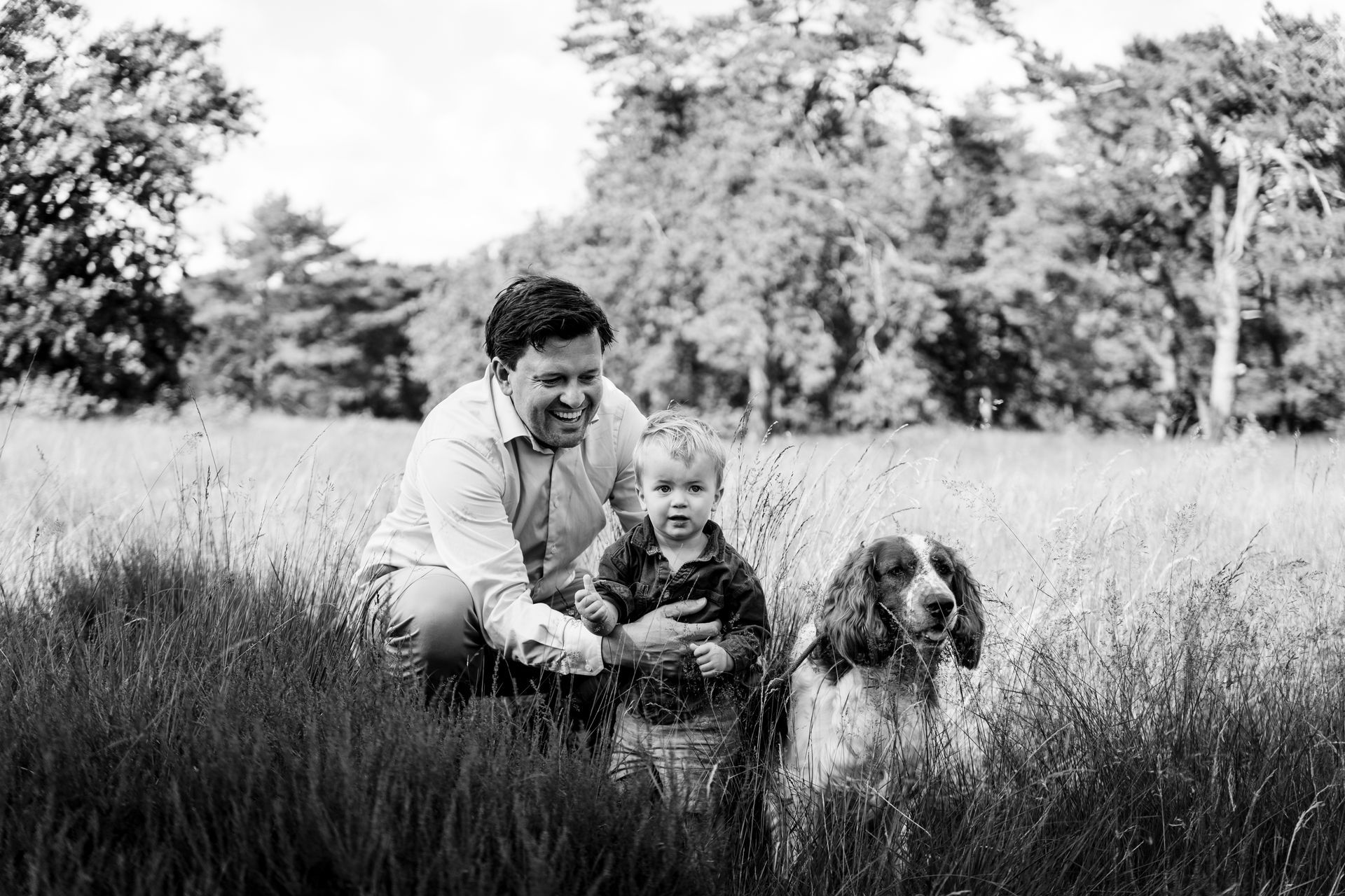 Een man en een kind hurken in het hoge gras met een hond in een veld; bomen op de achtergrond. Zwart-wit.