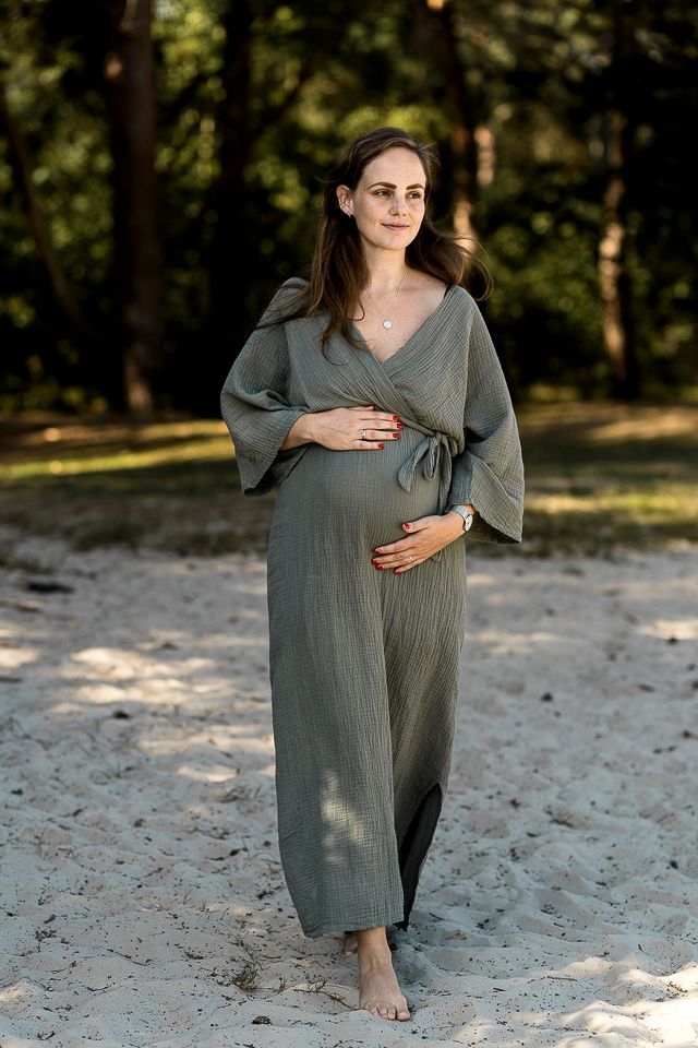 Een zwangere vrouw in een olijfgroene jurk staat op een zandstrand, met haar handen op haar buik.