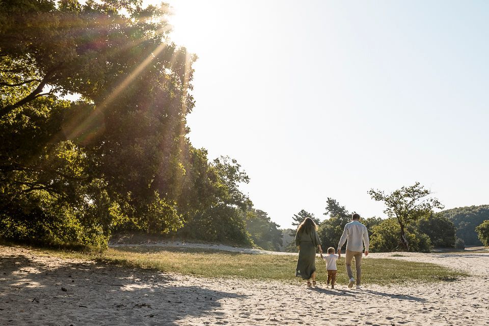 Een gezin loopt hand in hand over een zandpad richting de zon, met bomen aan de linkerkant.