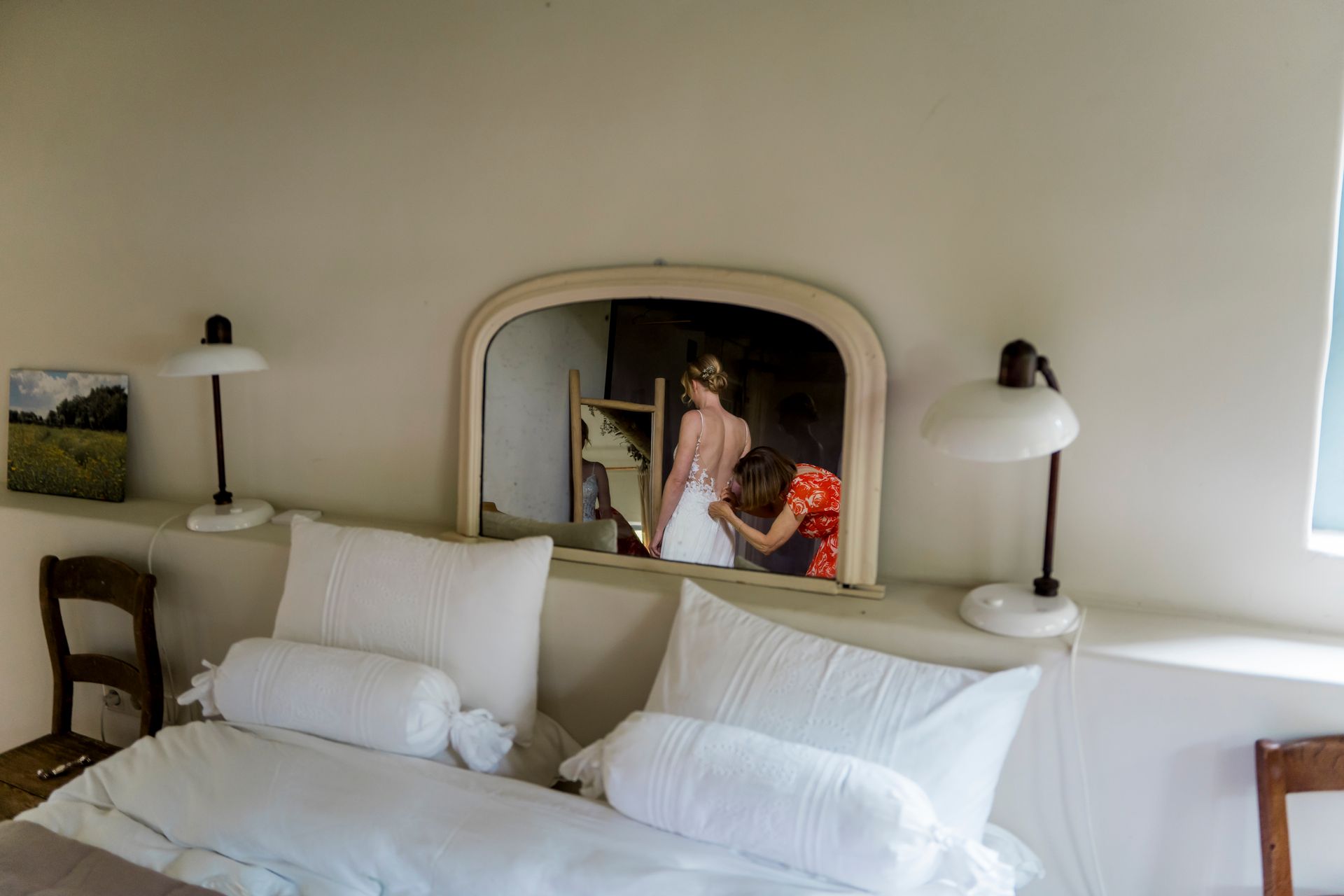 Slaapkamer met twee bedden, een spiegel waarin een vrouw te zien is die geholpen wordt bij het aantrekken van een jurk, en nachtlampjes.