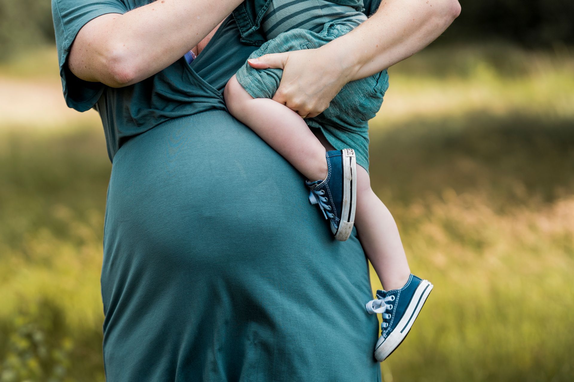 Een zwangere vrouw in een turquoise jurk houdt een klein kindje vast, dat een groen shirtje en blauwe schoenen draagt, in de buitenlucht.