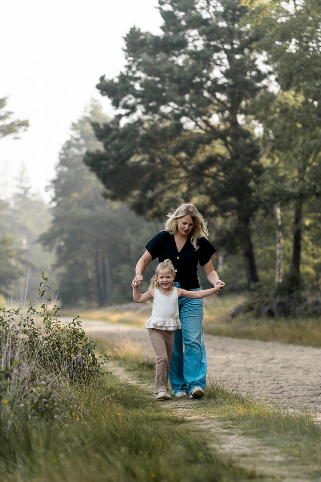 Vrouw en kind lopen hand in hand over een pad in een bos; beiden glimlachen.