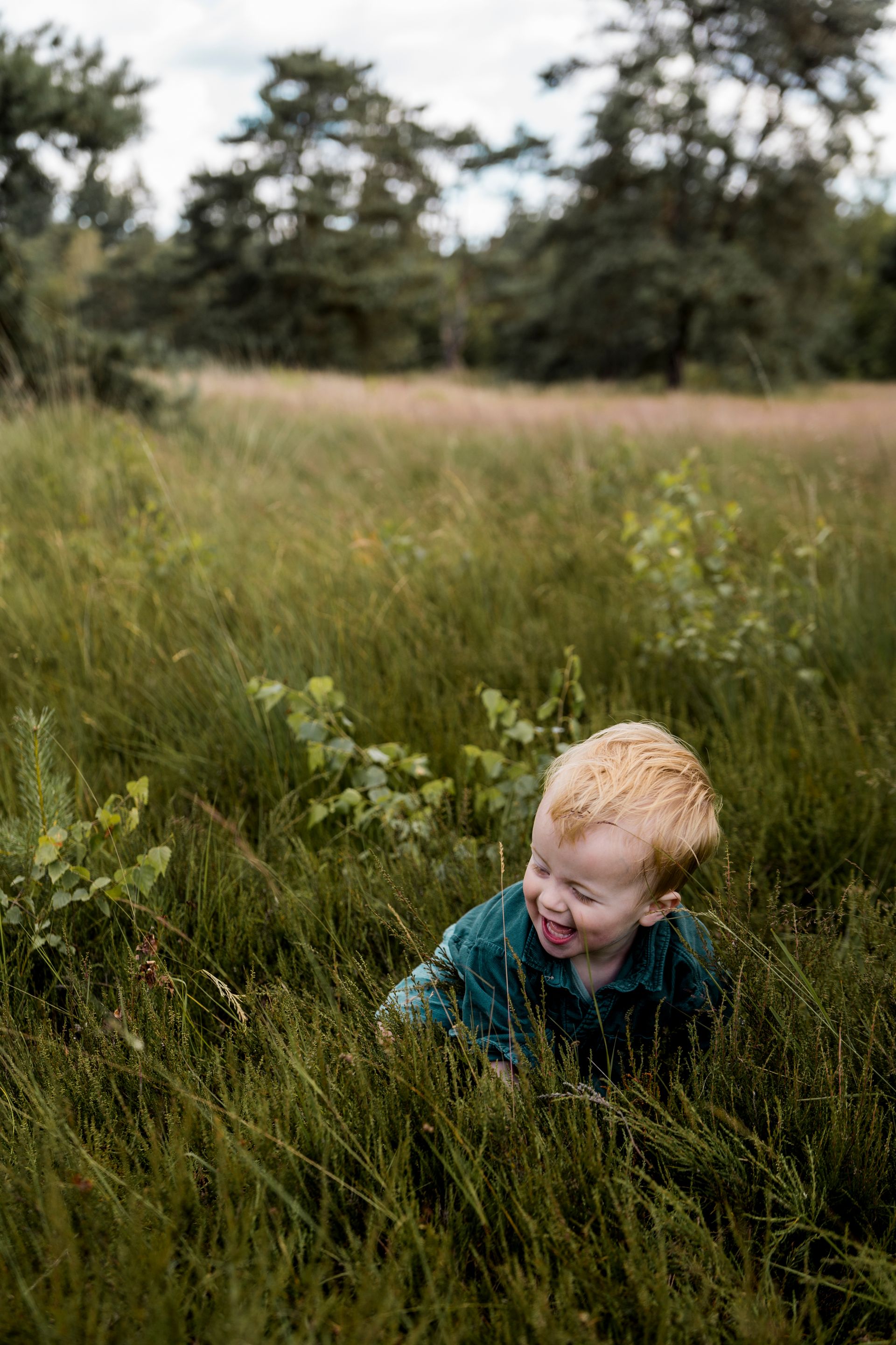 Blond kind speelt in het hoge groene gras, steekt zijn tong uit, met bomen op de achtergrond.