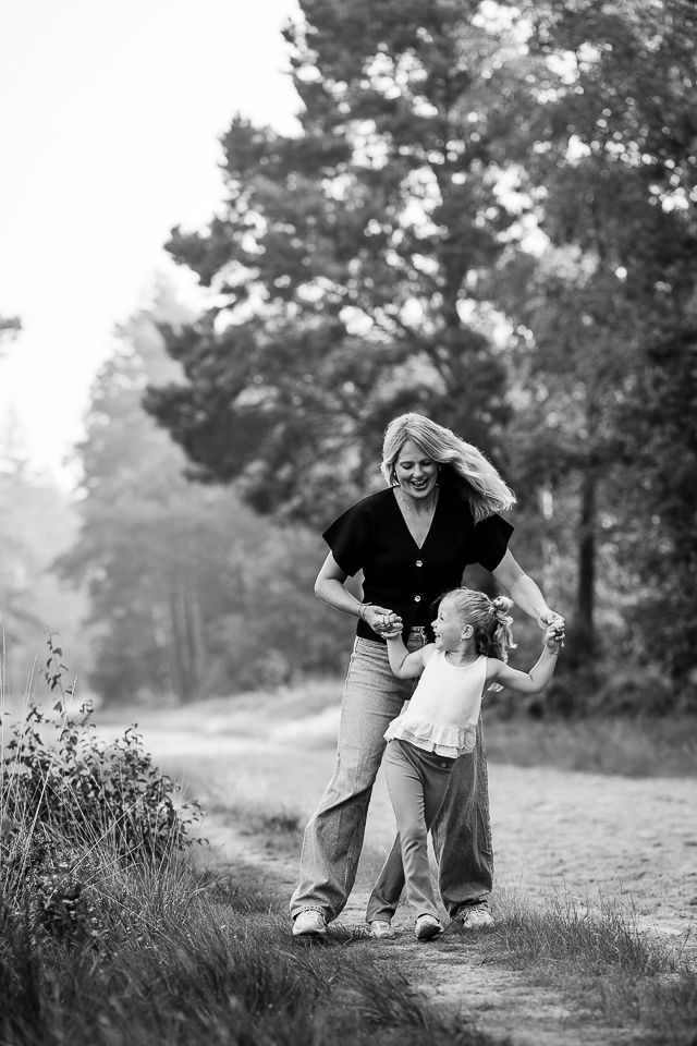 Vrouw en kind dansen op een pad in een bos; de vrouw heeft haar armen omhoog, het kind houdt haar handen vast. Zwart-wit.
