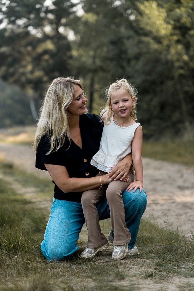 Een vrouw knielt en omhelst een kind op een zandpad. Ze glimlachen. Op de achtergrond zijn groene bomen te zien.