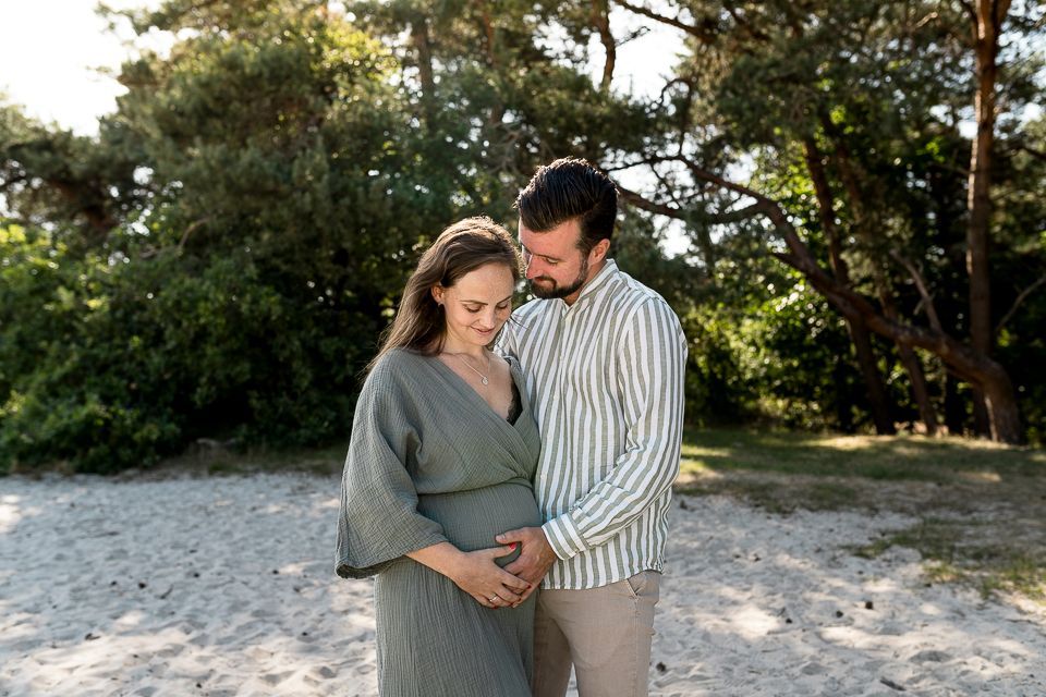 Een zwangere vrouw en haar partner houden hun buik vast en glimlachen op een strand voor de bomen.