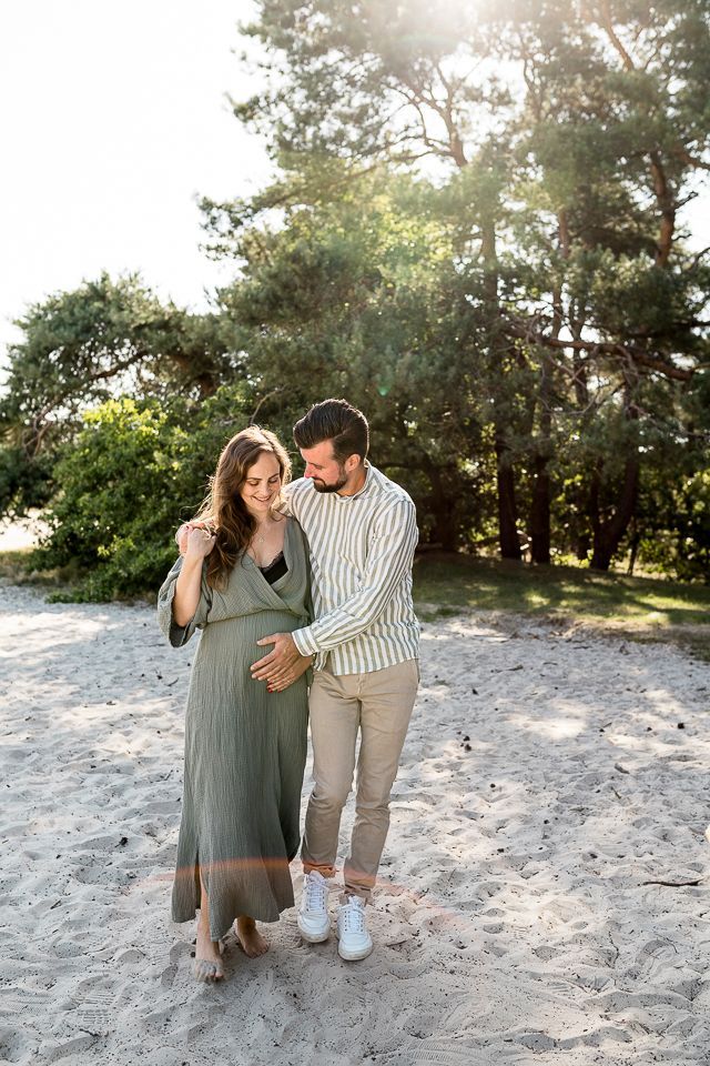Een zwangere vrouw en haar partner raken haar buik aan terwijl ze over het zand lopen, voor een bos.