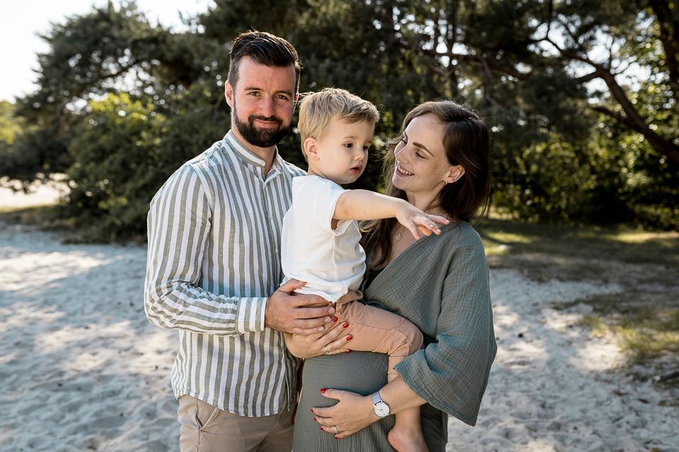 Een gezin van drie lacht buiten: een man houdt een kind vast, een vrouw kijkt toe.