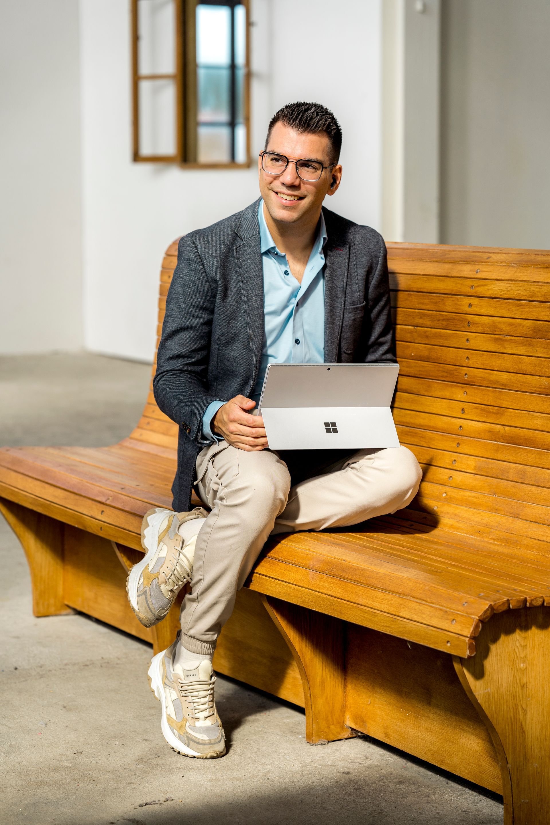 Een man zit op een houten bankje, werkt met zijn laptop en glimlacht. Hij draagt ​​een blazer, een blauw shirt, een beige broek en sneakers.