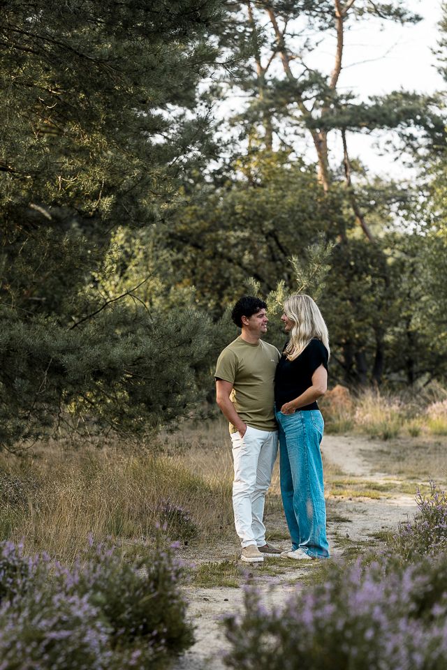 Een stel staat op een pad in het bos en kijkt elkaar aan. De man draagt ​​een groen shirt en een witte broek, de vrouw een zwart shirt en een blauwe spijkerbroek.