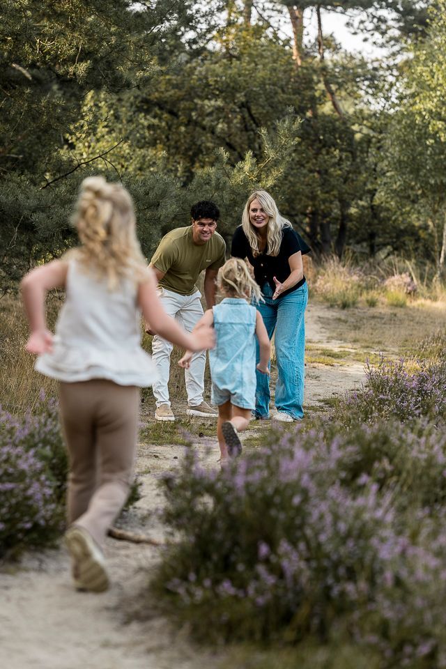 Een gezin speelt op een pad in het bos: twee jonge kinderen rennen naar volwassenen toe, paarse bloemen op de voorgrond.