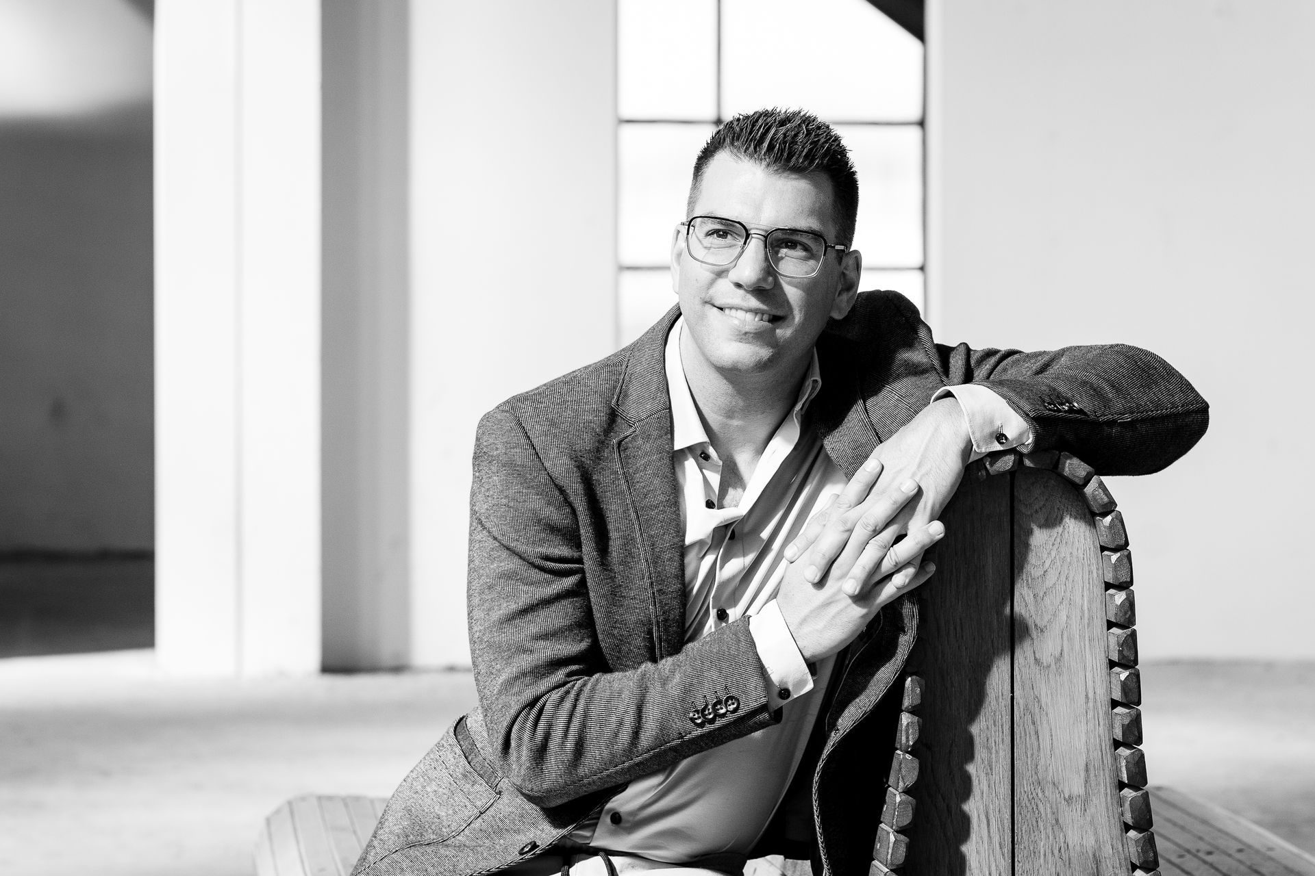 Een man met bril en blazer leunt glimlachend tegen een houten stoel in een moderne ruimte.