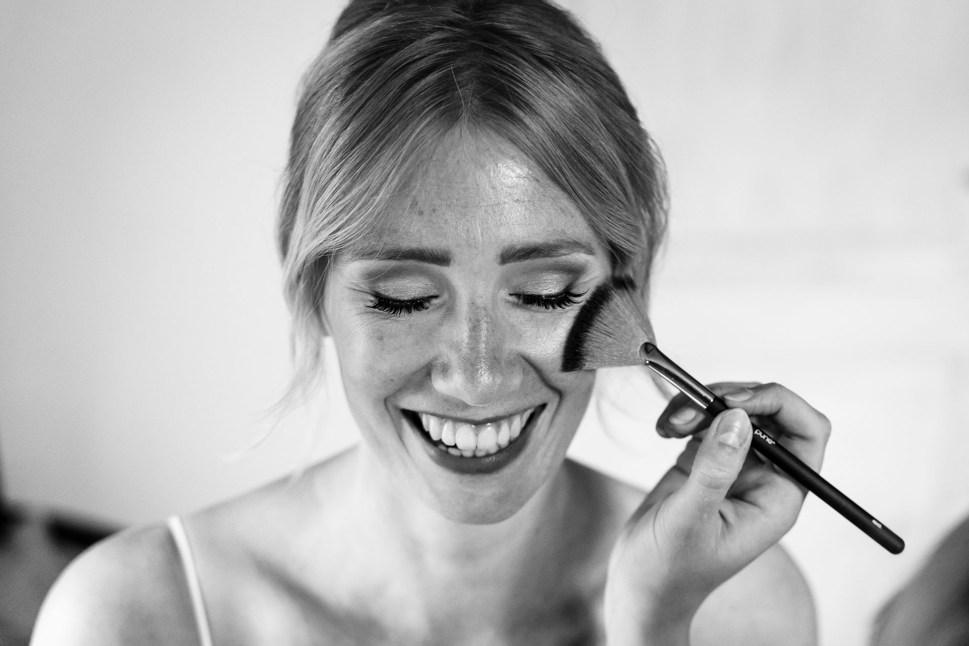 Vrouw glimlacht terwijl ze met een make-upkwast product op haar gezicht aanbrengt. Zwart-wit.