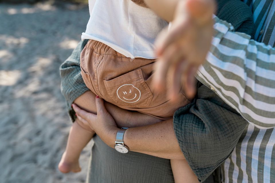 Persoon houdt een baby vast; de baby draagt een bruine korte broek met een lachend gezichtje.