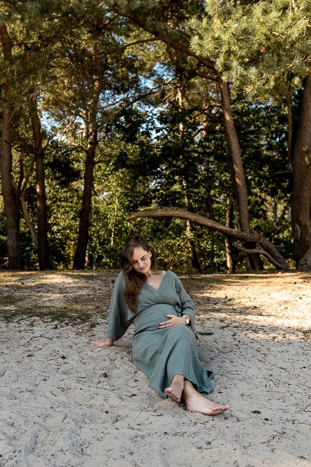 Een zwangere vrouw zit in het zand, gekleed in een turquoise jurk, in een bosrijke omgeving en raakt haar buik aan.