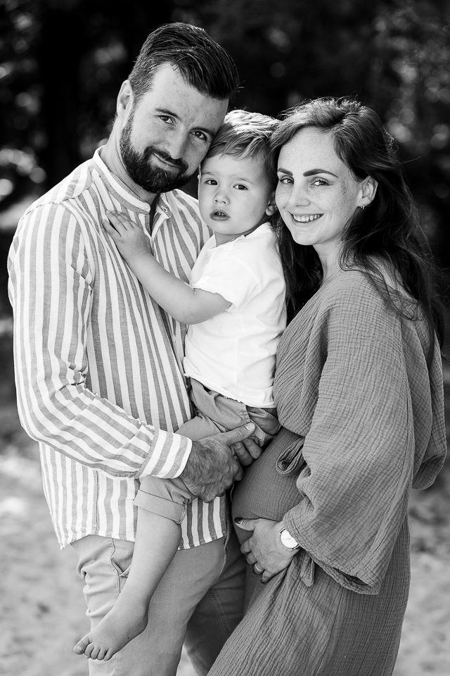 Familieportret: vader met kind, moeder zwanger, allen lachend. Buiten, zwart-wit.