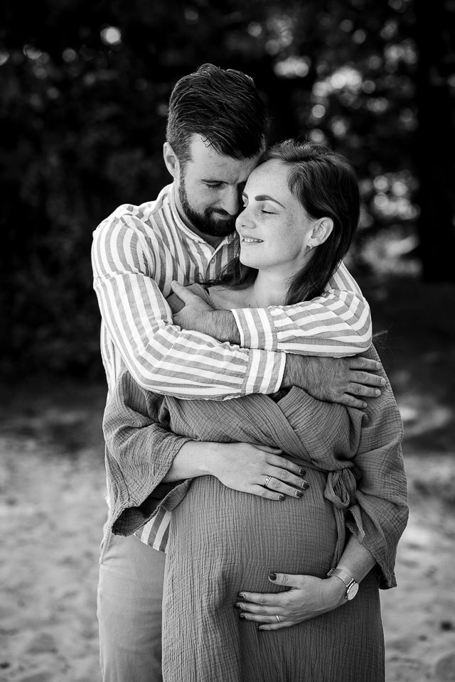 Een man omhelst een zwangere vrouw in de buitenlucht. Beiden glimlachen, de man heeft zijn armen om haar heen geslagen en zijn handen rusten op haar babybuik. Zwart-wit.