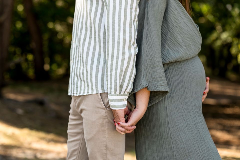 Een man en een zwangere vrouw houden elkaars hand vast, buiten. Op de achtergrond een bos.