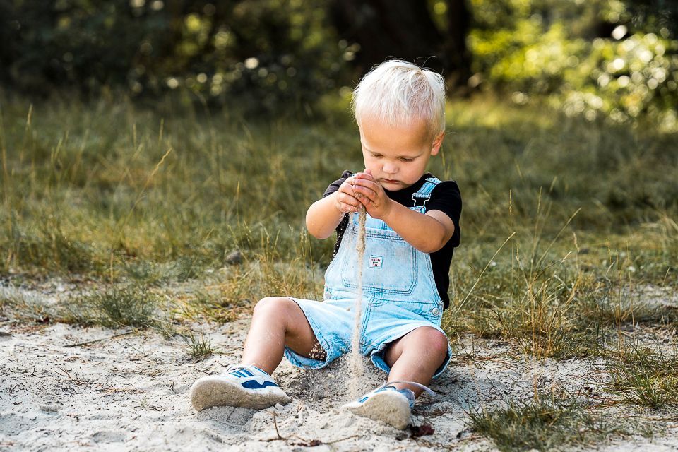 Een blond peutertje in een tuinbroek zit in het zand en bekijkt een stokje voor een grasveld.