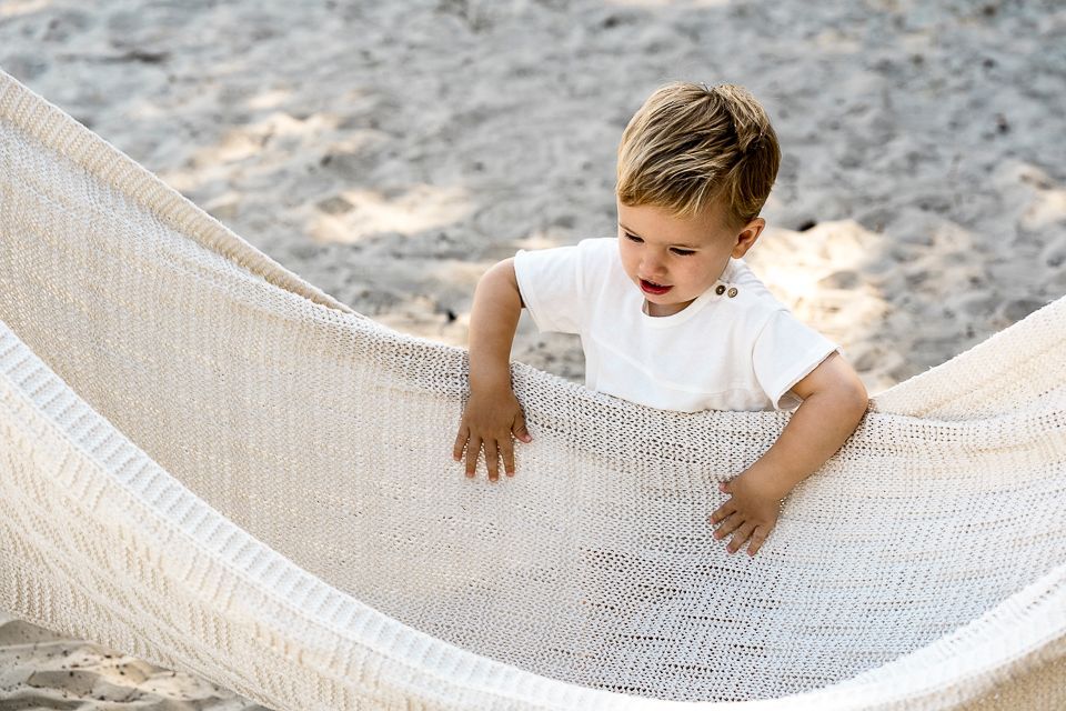 Een jongen in een wit shirt speelt in een crèmekleurige hangmat op een zandstrand.