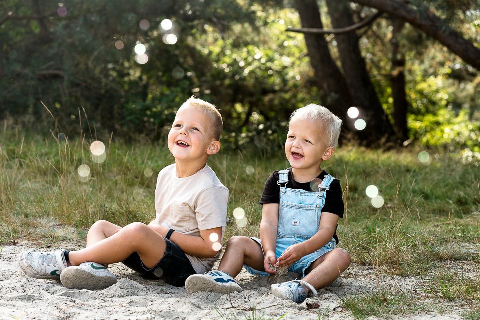 Twee jonge kinderen zitten buiten en kijken lachend naar de bubbels, een van hen draagt een overall.