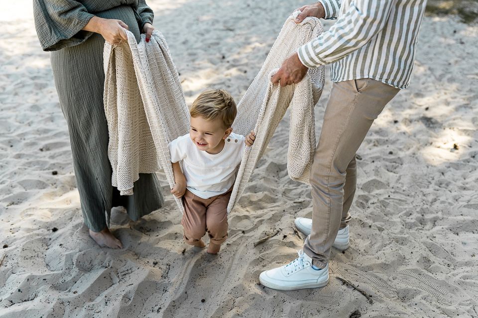 Ouders die een deken vasthouden en een peuter optillen op een zandstrand.
