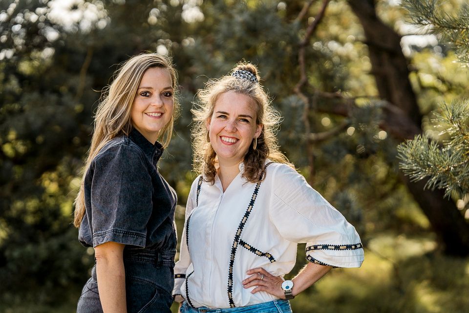 Twee lachende vrouwen poseren buiten bij bomen. De ene draagt een spijkerbroek, de andere een witte top met zwarte bies.