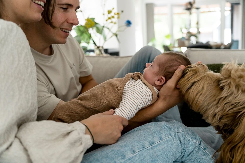 Een stel met een pasgeboren baby en een hond, die allemaal samen op de bank in de woonkamer zitten.