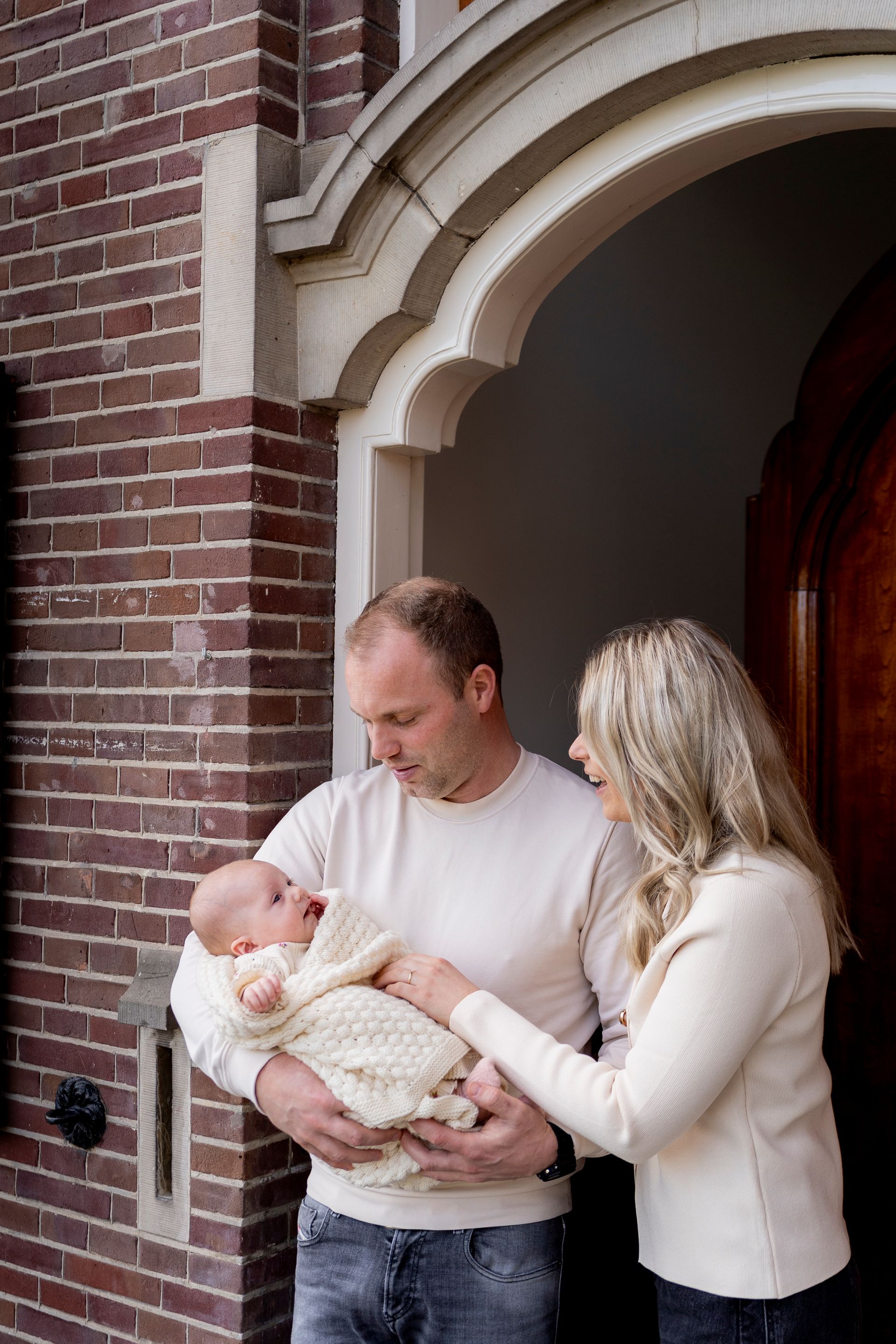 Een man houdt een ingewikkelde baby vast, terwijl een vrouw toekijkt, bij de ingang van een bakstenen gebouw.