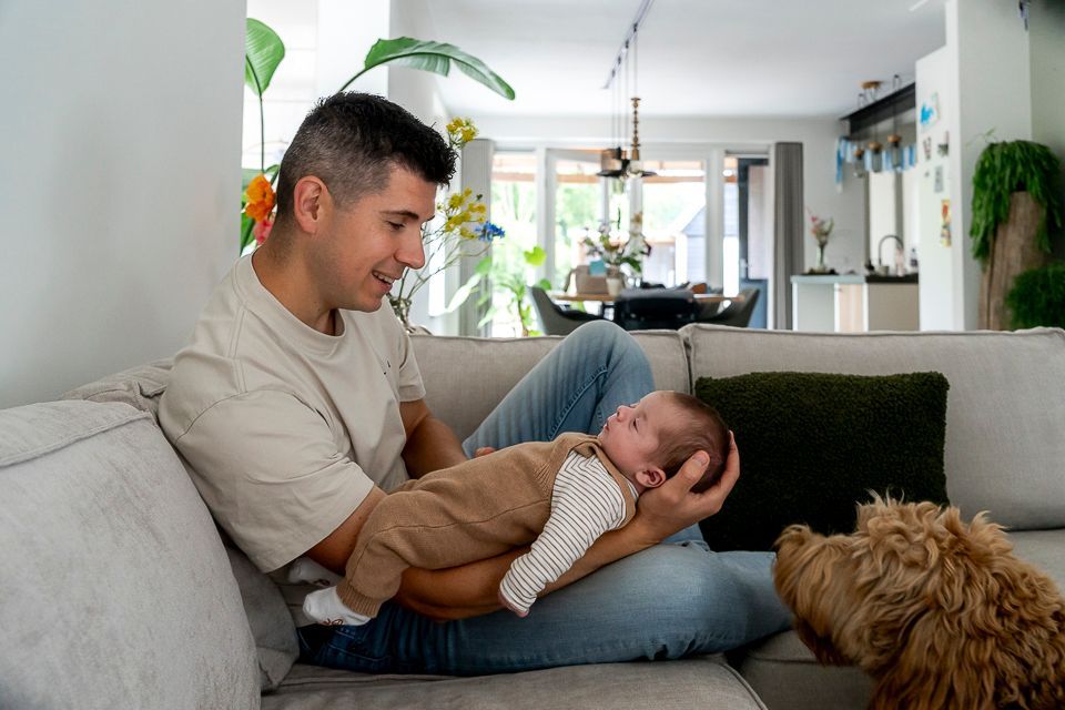 Een man houdt een baby vast op een bank en kijkt naar het kind. Een hond kijkt toe. Woonkamer.