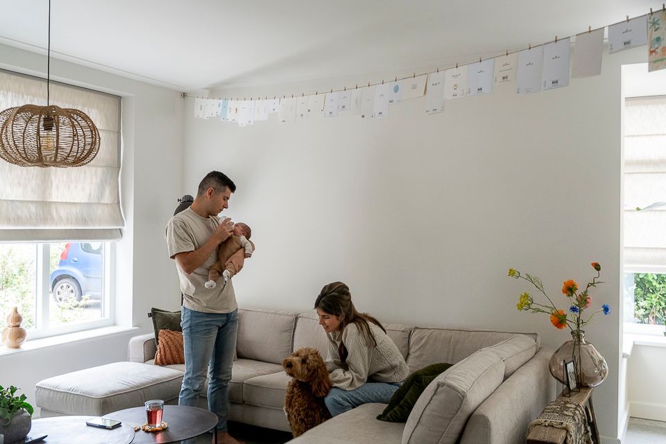 Gezin met pasgeborene in de woonkamer; ouders, hond en een slinger van kunstwerken.