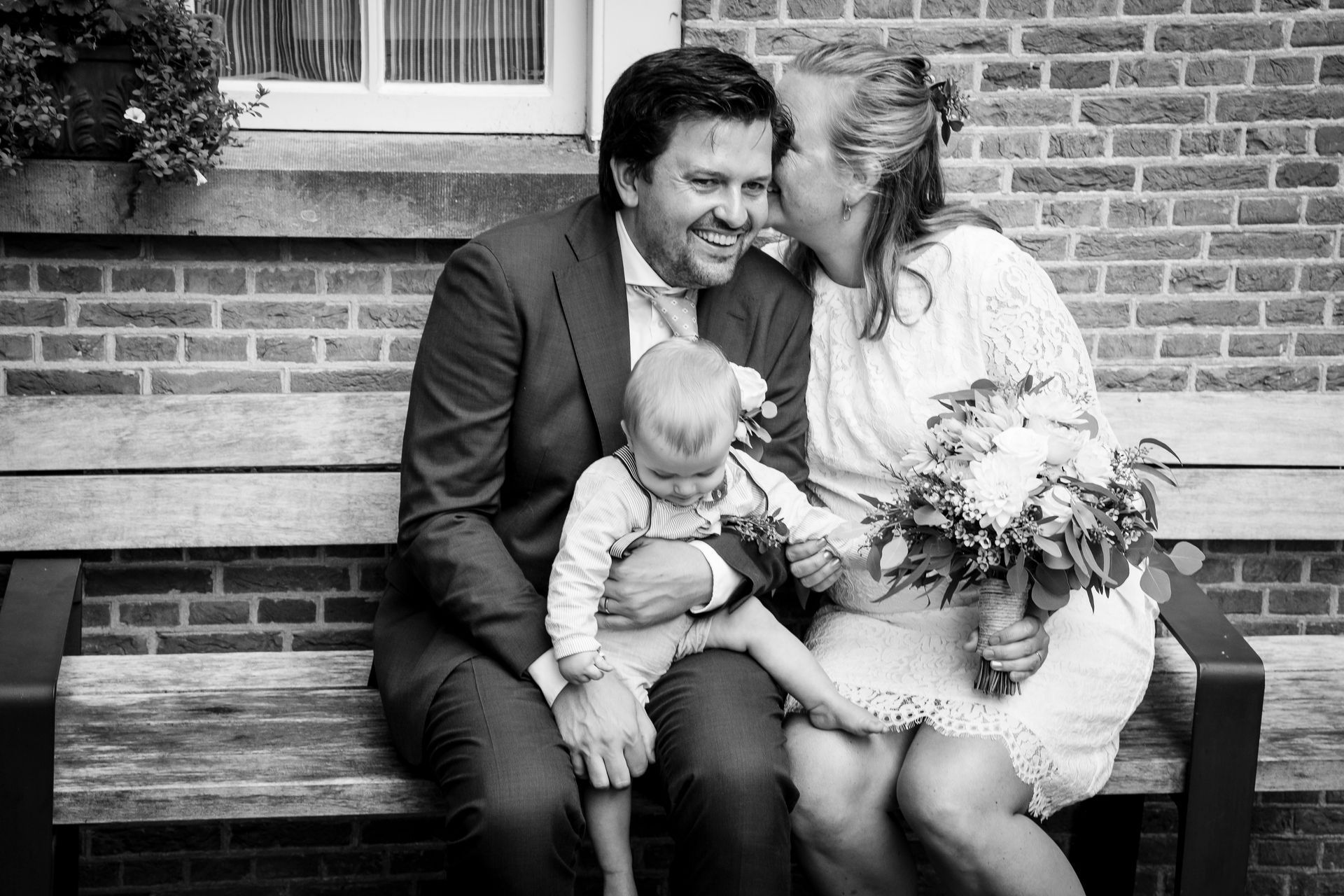 Familieportret: Een echtpaar lacht en houdt een kind vast, zittend op een bankje voor een bakstenen gebouw.