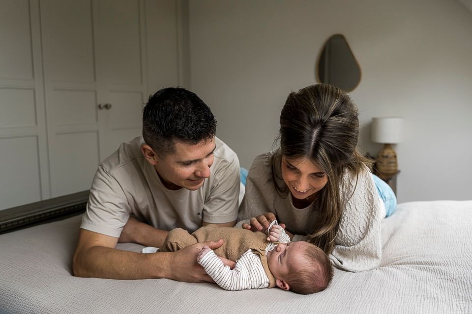 Ouders kijken naar hun pasgeboren baby in bed. Ze bevinden zich in een lichte slaapkamer.