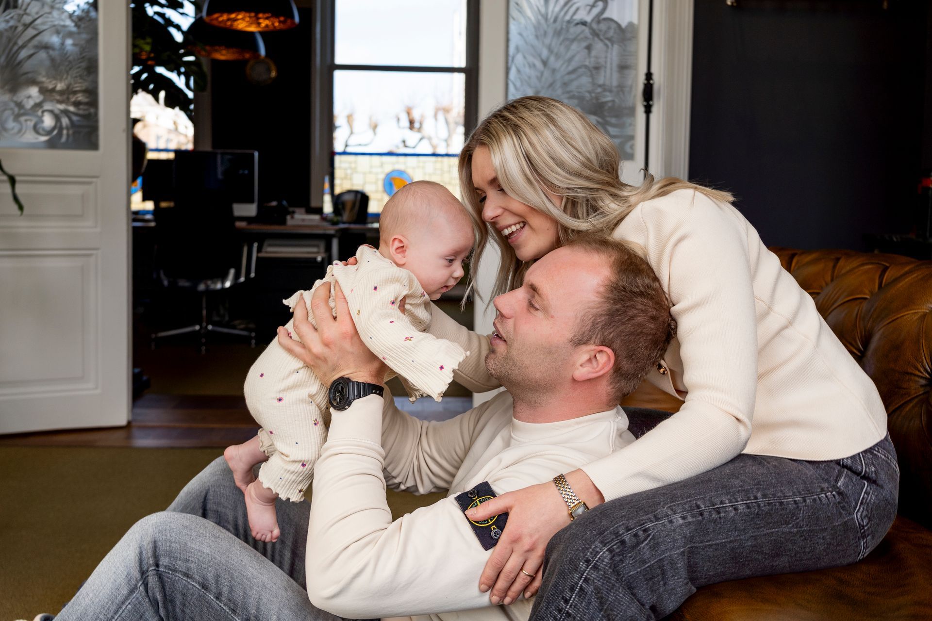 Een gezin van drie lacht en geniet van elkaars gezelschap. De vader houdt de baby omhoog, de moeder buigt zich voorover. Binnen, warme kleuren.