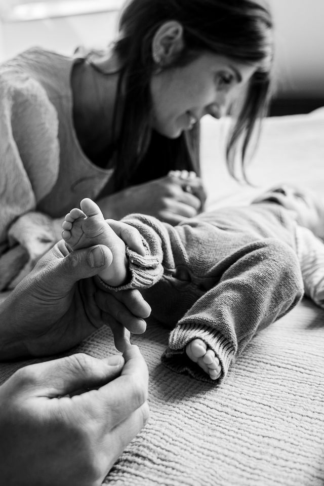 Iemand raakt zachtjes de voetjes van een baby aan op een bed. De moeder kijkt toe met een tedere uitdrukking. Zwart-wit.