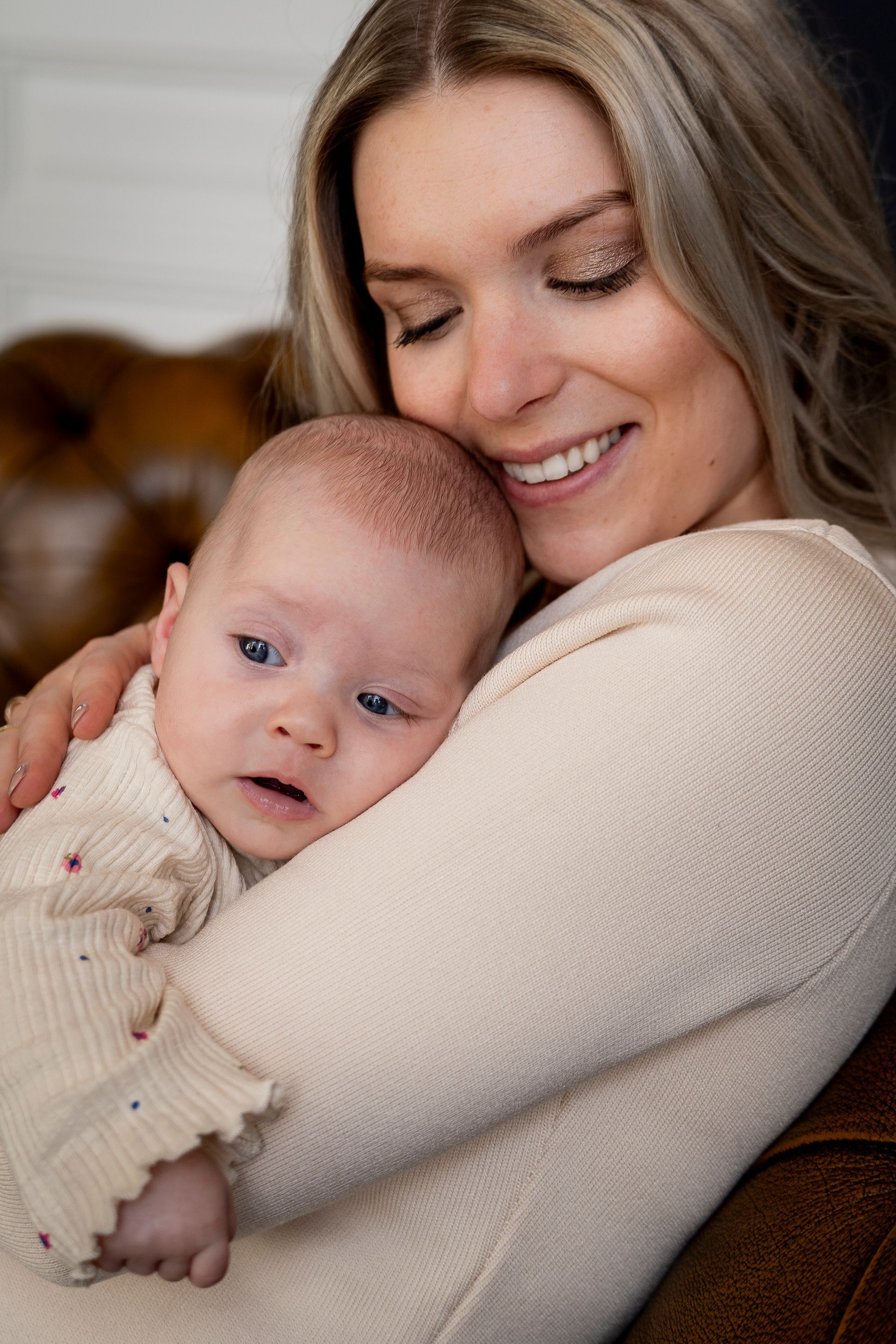 Een vrouw lacht en houdt haar baby dicht tegen zich aan. Beiden dragen lichtgekleurde truien.