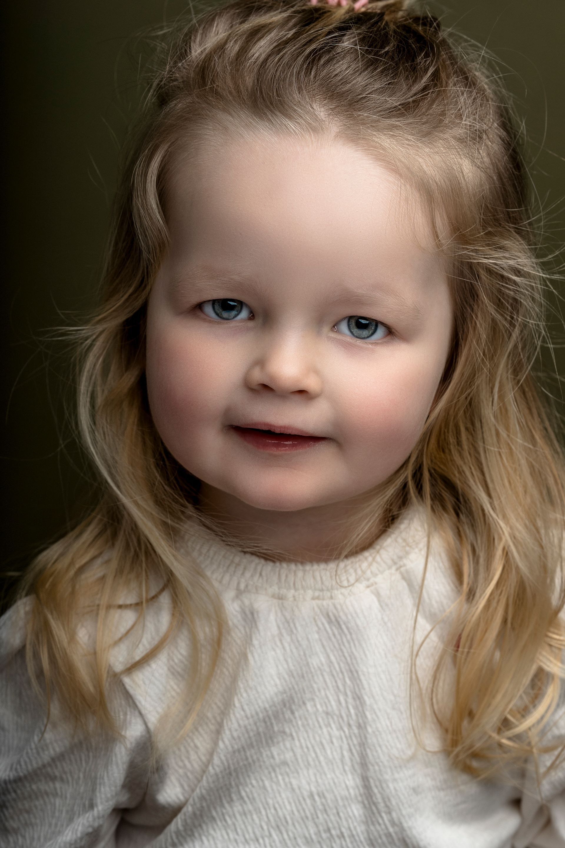 Een blond kind met blauwe ogen, dat lichtjes glimlacht, draagt een crèmekleurige trui en staat afgebeeld tegen een donkergroene achtergrond.