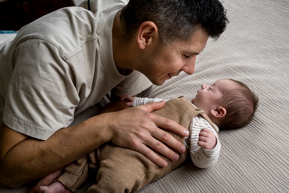 Een man kijkt naar een baby die op een bed ligt. De handen van de man rusten zachtjes op de romp van de baby. Beiden glimlachen.