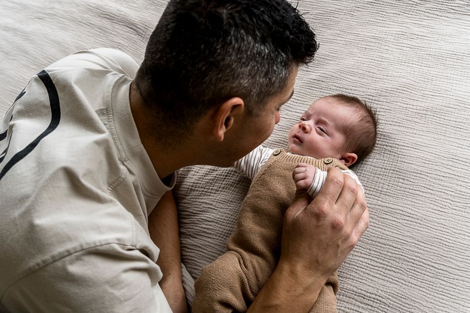 Een man kijkt naar een pasgeboren baby die op een deken ligt. De baby draagt een bruine overall.