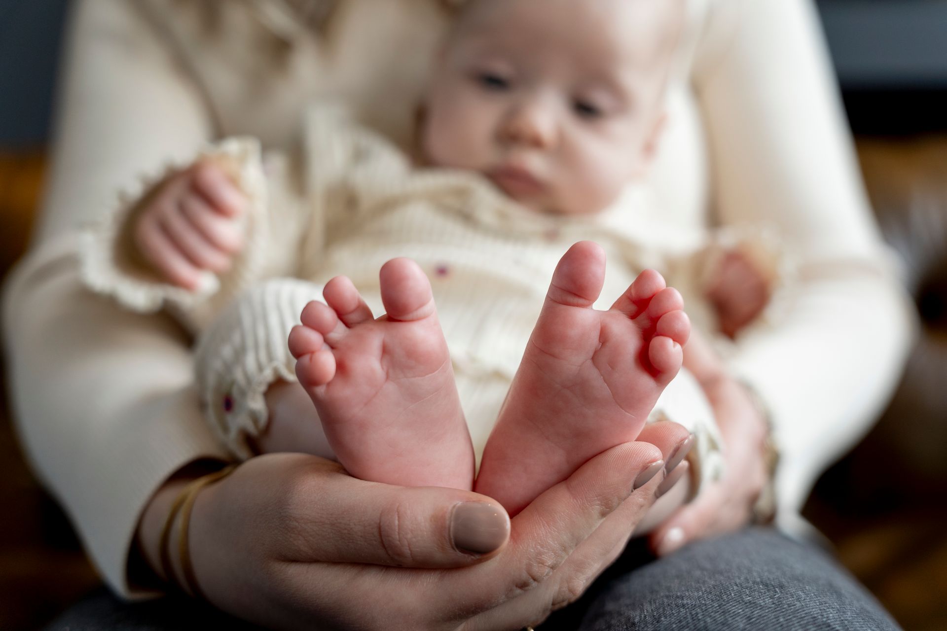 De voetjes van de baby zijn scherp in beeld, vastgehouden door een persoon. De baby draagt ​​crèmekleurige kleding.
