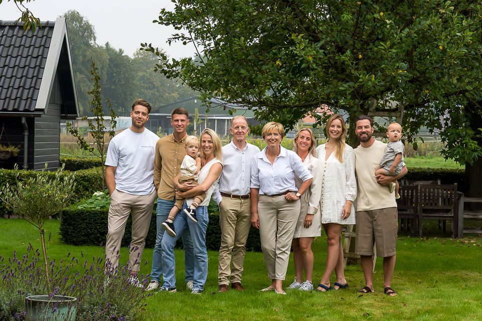 Een gezin van elf personen poseert lachend buiten in een tuin. Volwassenen en kinderen staan ​​op het gras; op de achtergrond zijn bomen en een huis te zien.