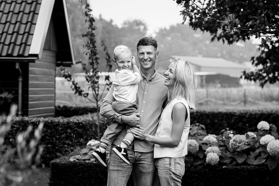 Een gezin van drie lacht buiten, een man houdt een kind vast. Een vrouw lacht ernaast. Zwart-wit.