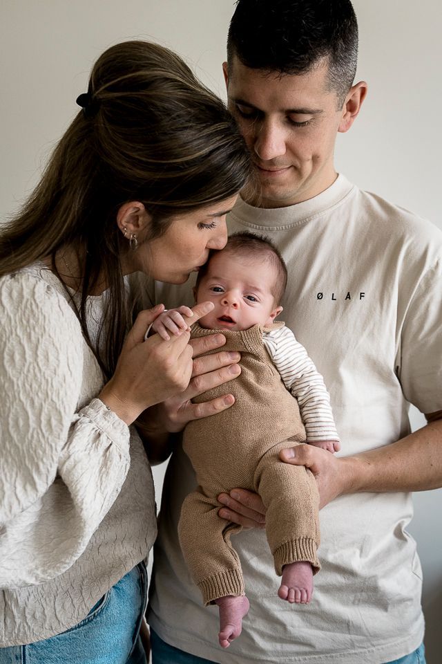 Ouders die elkaar kussen en een baby vasthouden. De baby draagt een beige rompertje. Neutrale achtergrond.